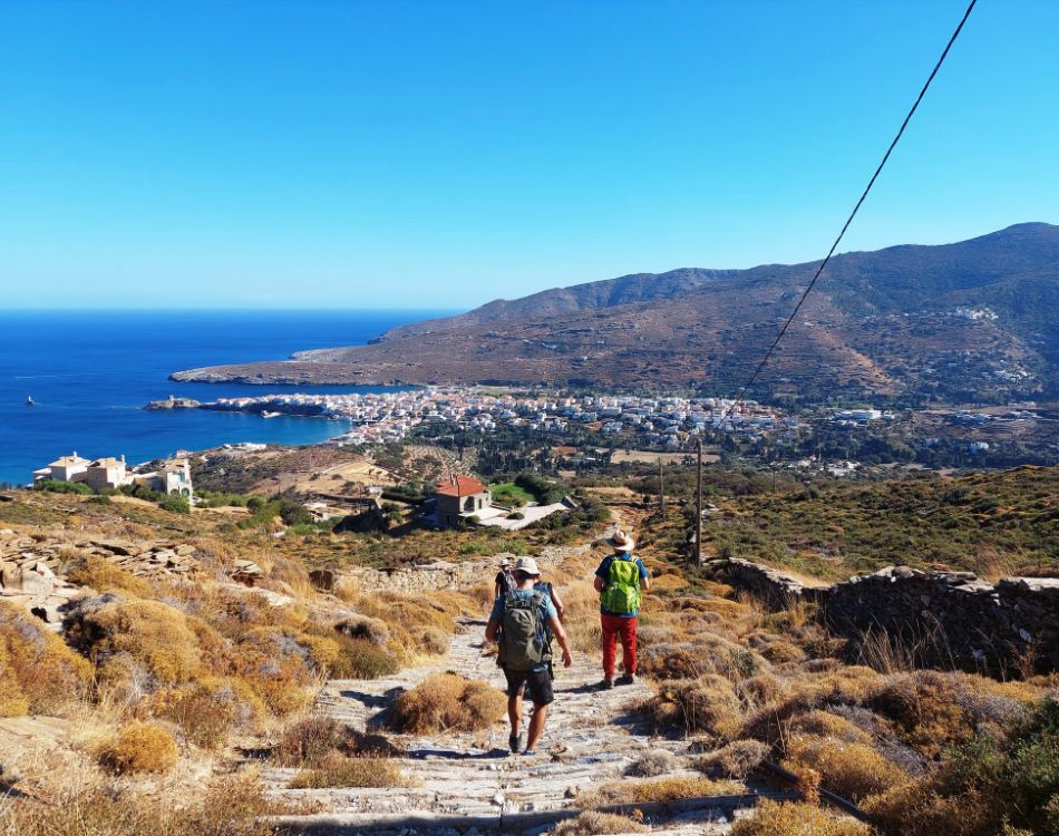 Two hikers walking down a rocky trail towards a coastal village on Andros island, Greece, with a stunning view of the blue sea and mountains in the background.