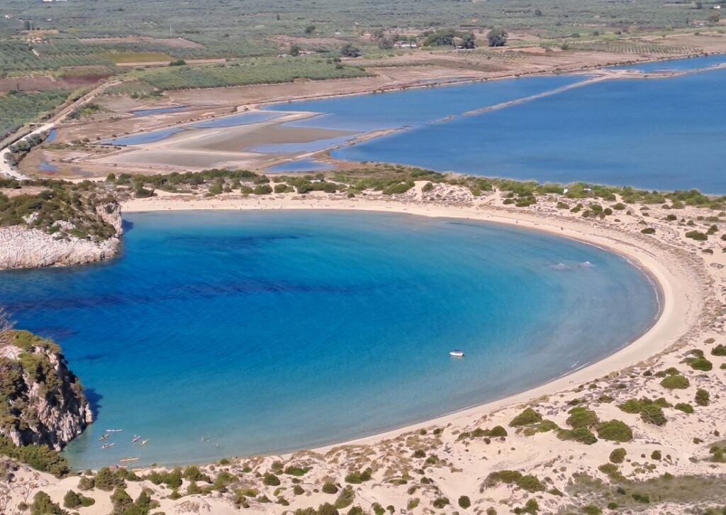 Aerial view of the iconic Ω (Omega) shaped Voidokilia Beach and Gialova Lagoon, showcasing turquoise waters and golden sands in Greece.
