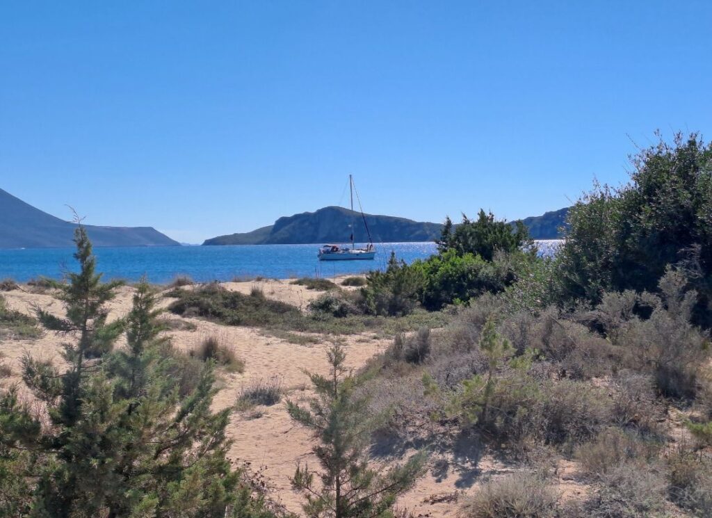 Sandy path leading to a quiet beach with a sailboat anchored in turquoise waters near Gialova Lagoon in the Peloponnese, Greece.