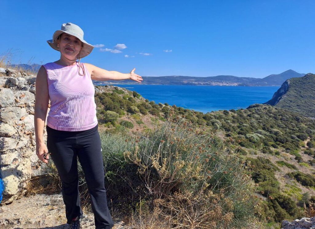 Evgenia of the Planet Greece pointing toward Voidokilia Beach and the Ionian Sea from a hillside viewpoint near Pylos, Greece.