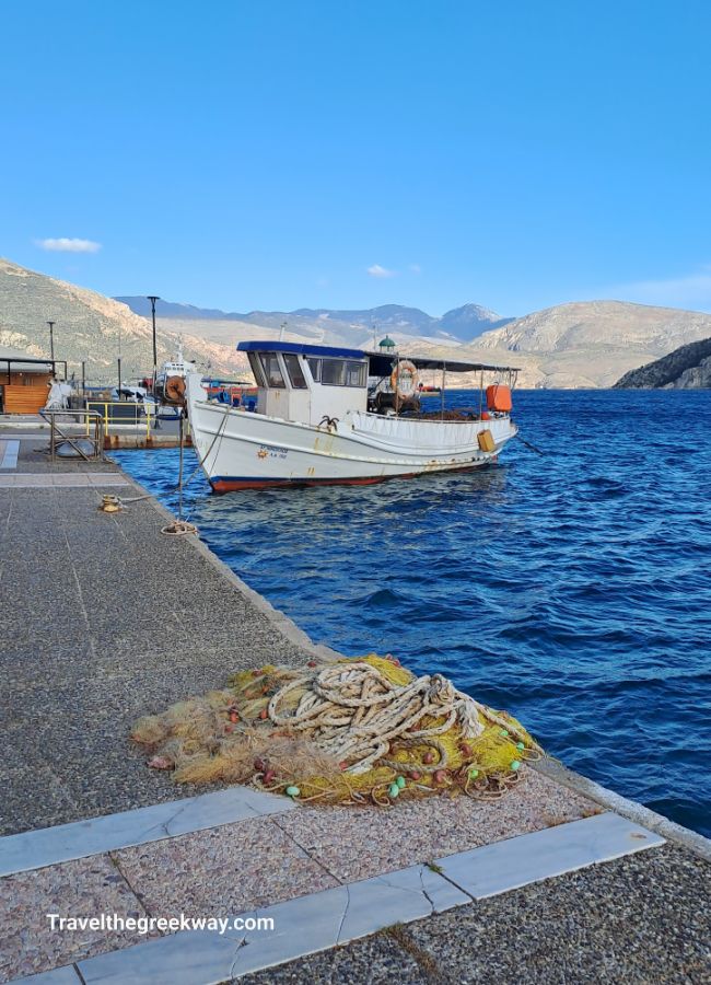 A fishing boat docked at a seafront pier with mountains in the background and fishing nets on the ground.