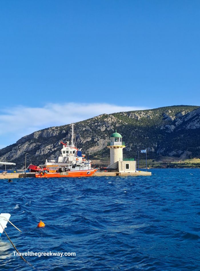 A lighthouse and boats at a pier with mountains in the background on a sunny day.