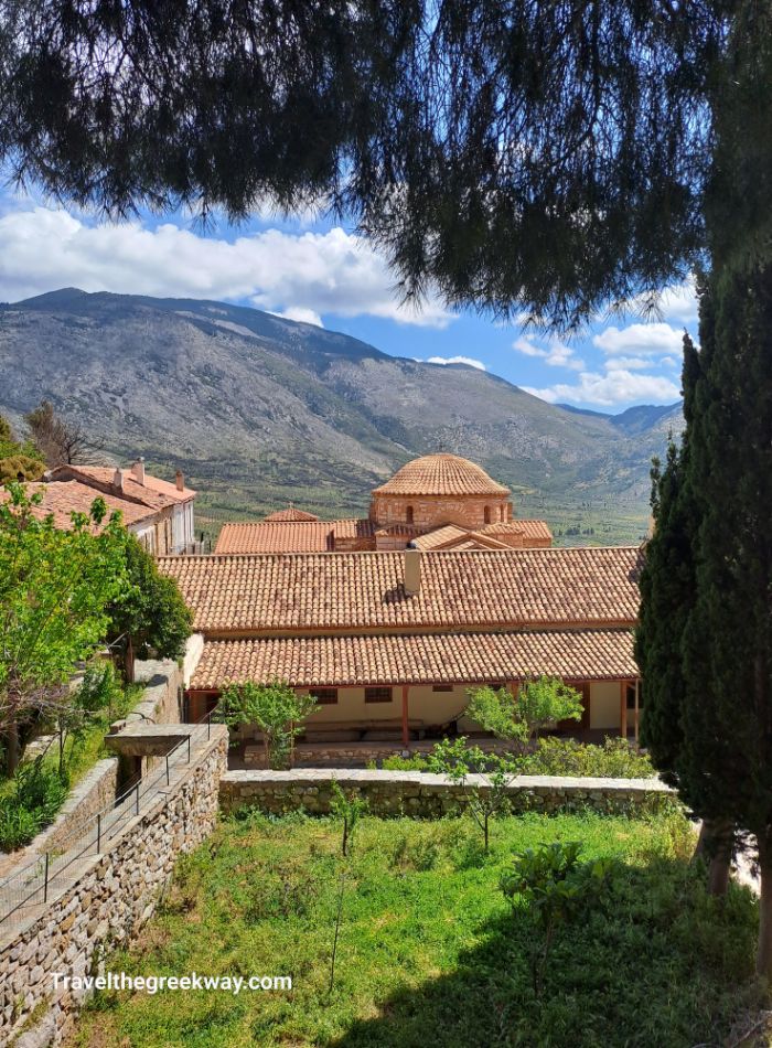 View of a Byzantine monastery with terracotta roofs nestled in a green valley, surrounded by mountains.