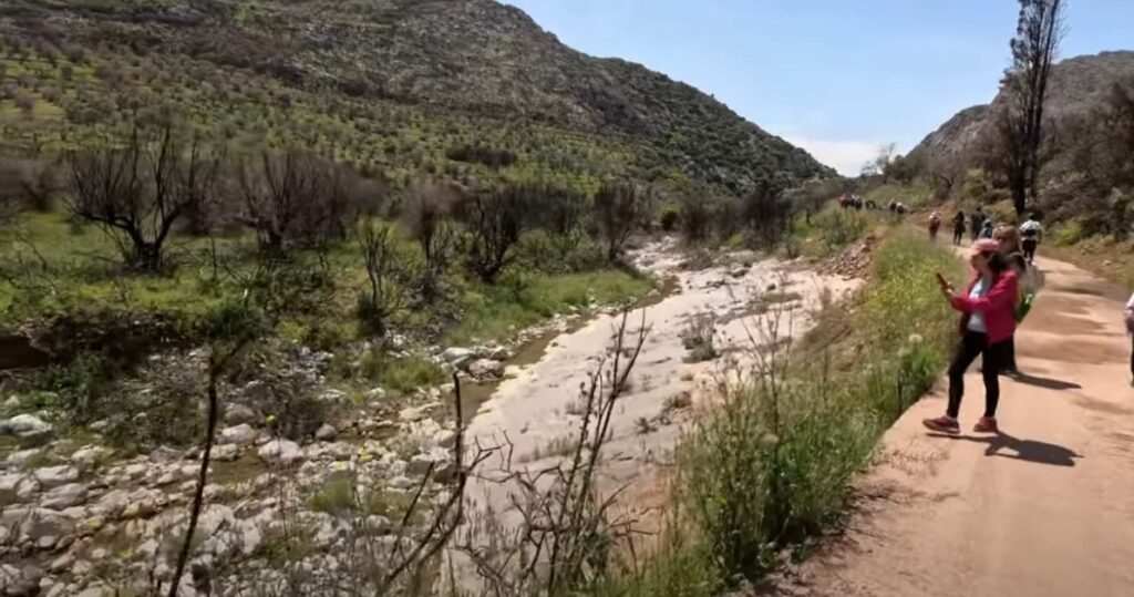 Hikers walking along a dirt path beside a stream with hills and greenery in the background.