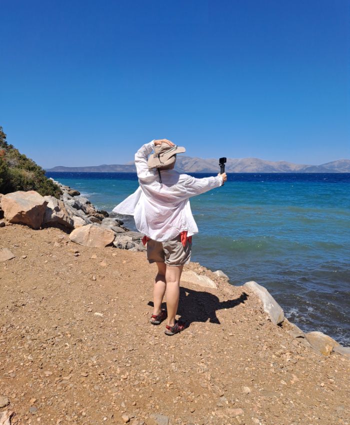 Person wearing a white shirt and hat, holding a camera, standing on a rocky shore with the sea and mountains in the background under a clear blue sky.