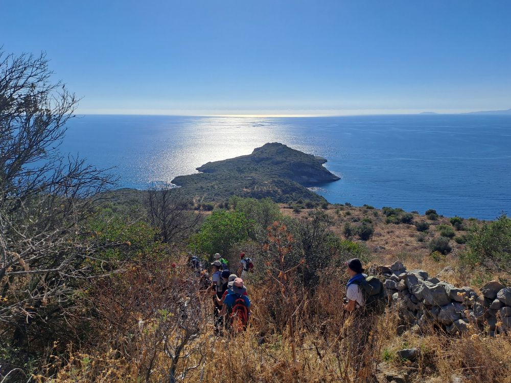 Hikers on a scenic trail in Mani, Peloponnese, overlooking the blue sea and a small island under a bright sky.