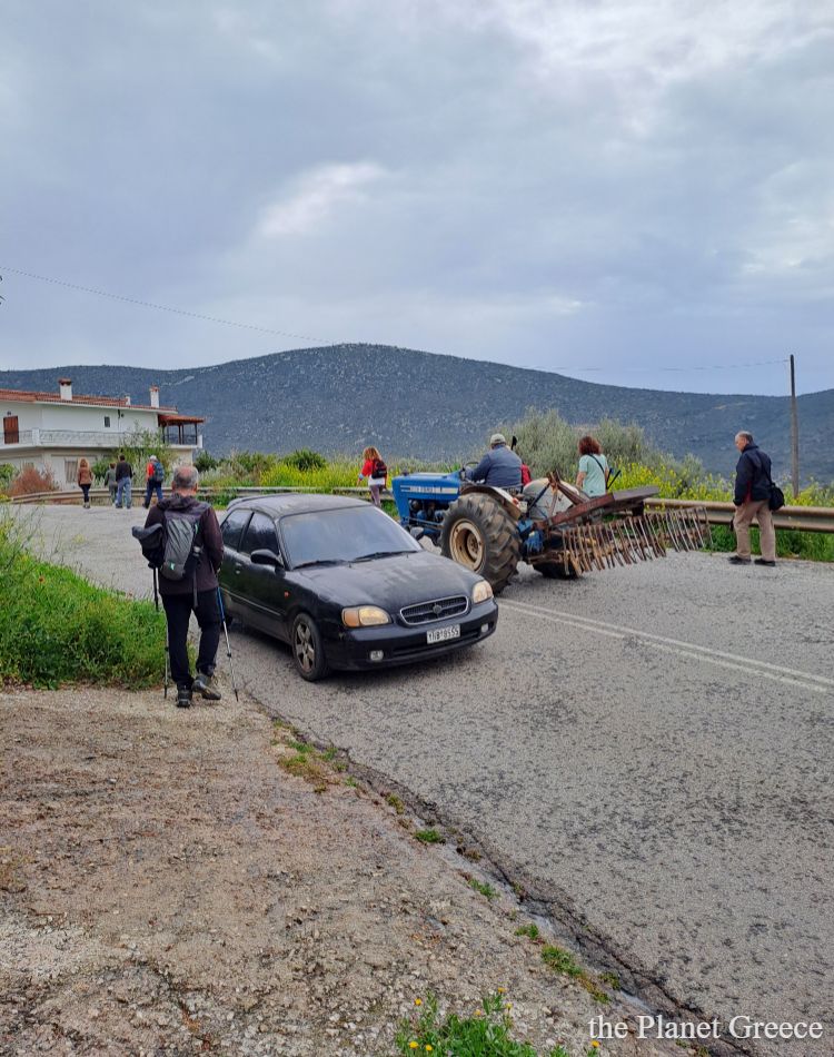 A rural road scene in the Peloponnese with hikers, a passing tractor, and a car, surrounded by green hills.