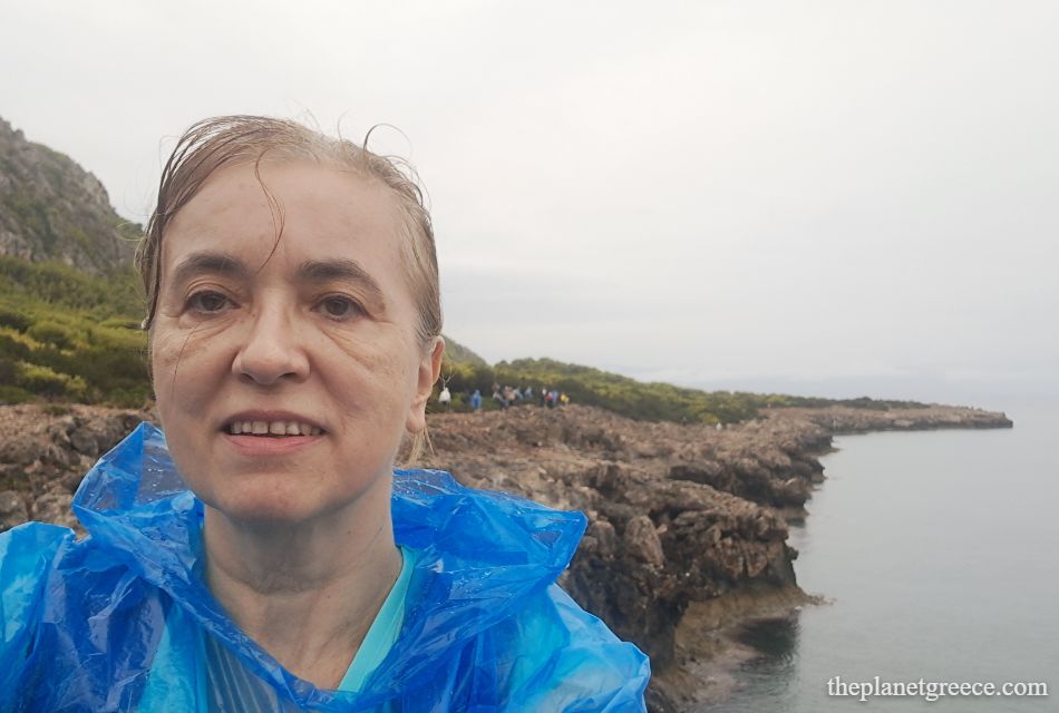 Evgenia in a blue raincoat on the rocky coastal trail while hiking to Melagkavi Lighthouse in Loutraki Greece.