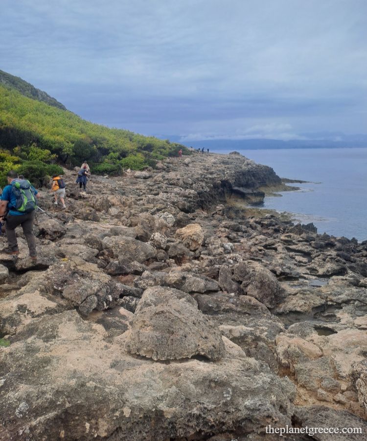 Hikers walking over rocky terrain on the coastal trail to Melagkavi Lighthouse in Loutraki Greece.