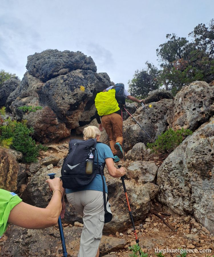Hikers climbing rocky path marked with yellow trail signs on the Melagkavi Lighthouse hike in Loutraki Greece.