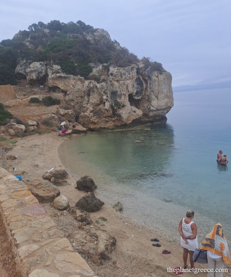 Small beach at Heraion in Loutraki, Greece, with swimmers and rocky cliffs in the background.